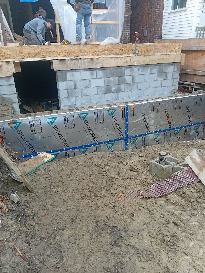 Construction site showing concrete foundation with blue protective barrier wrap and workers on temporary platform above the foundation