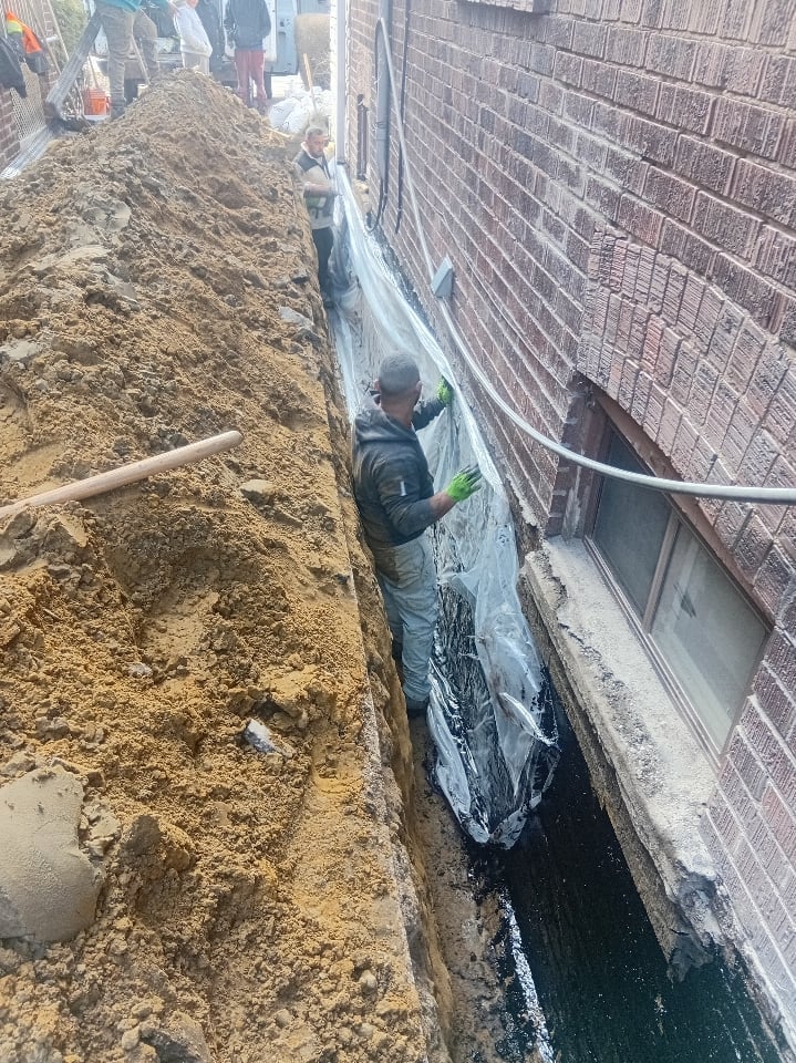 Worker in trench applying waterproof coating to brick foundation wall during construction