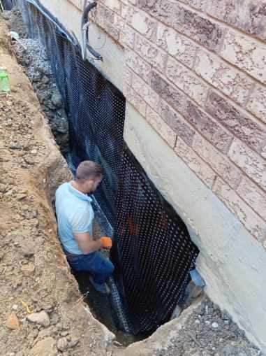 Man in light blue shirt installing black drainage matting against brick foundation of a building
