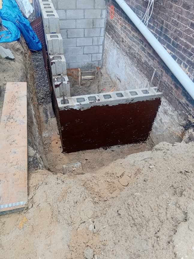 A deep excavated basement entrance hole in sandy soil between brick buildings with metal reinforcement at ground level