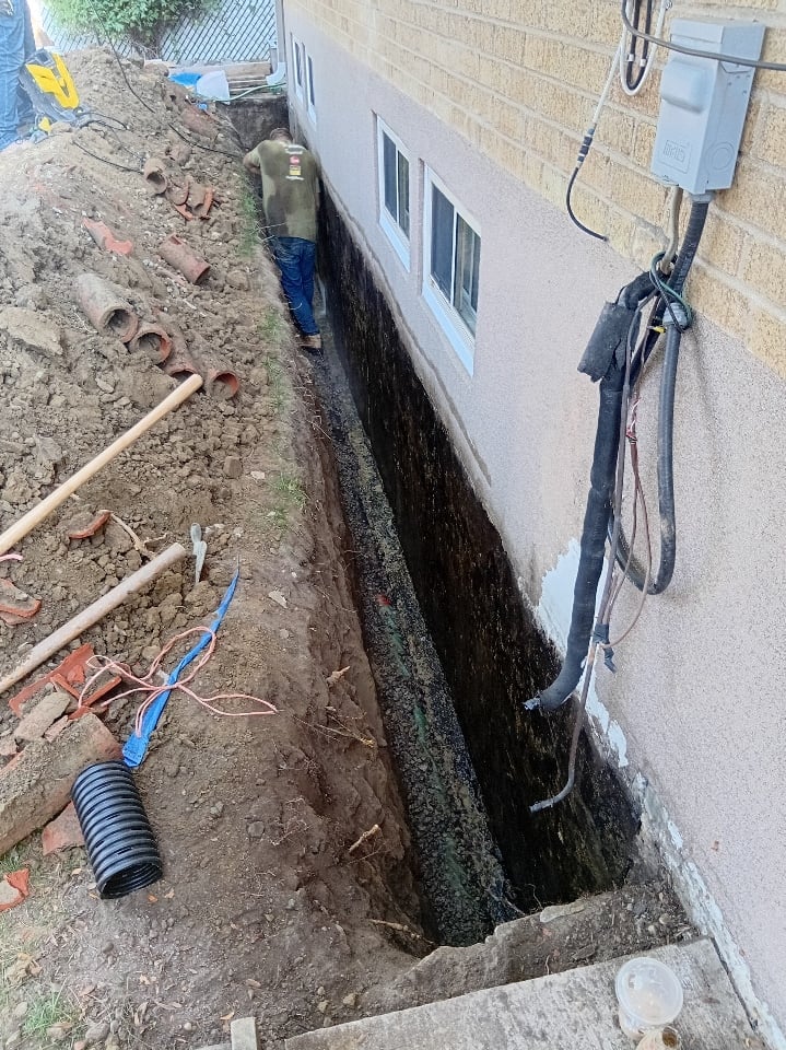 Worker in trench next to house foundation with exposed pipes and electrical conduit during construction or renovation work